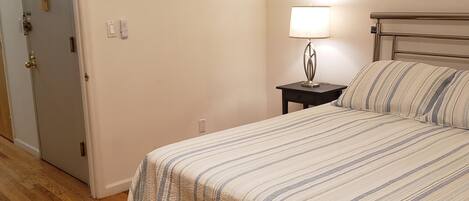 A bedroom with a bed, a nightstand, a lamp, a door, and a framed picture on the wall.