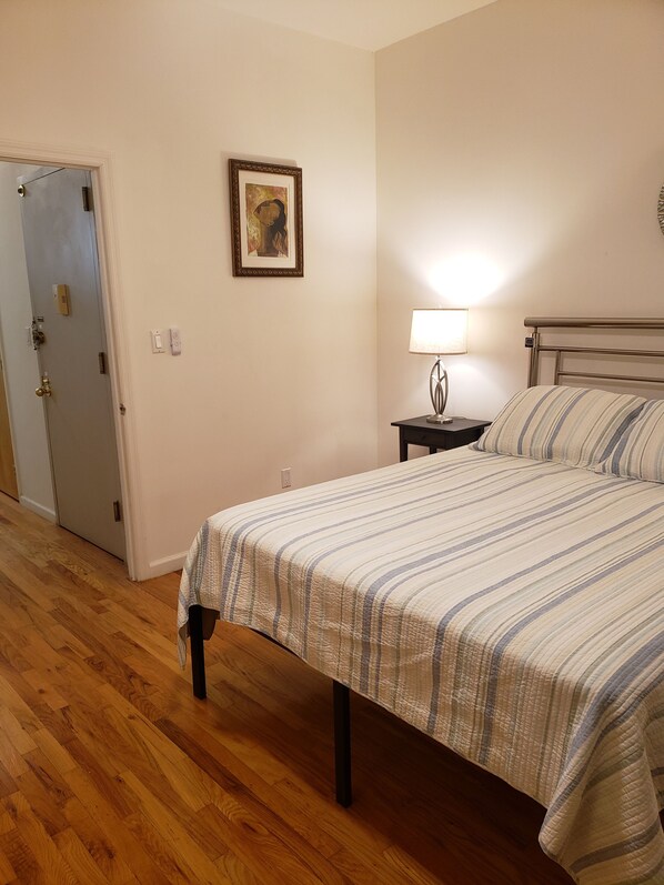 A bedroom with a bed, a nightstand, a lamp, a door, and a framed picture on the wall.