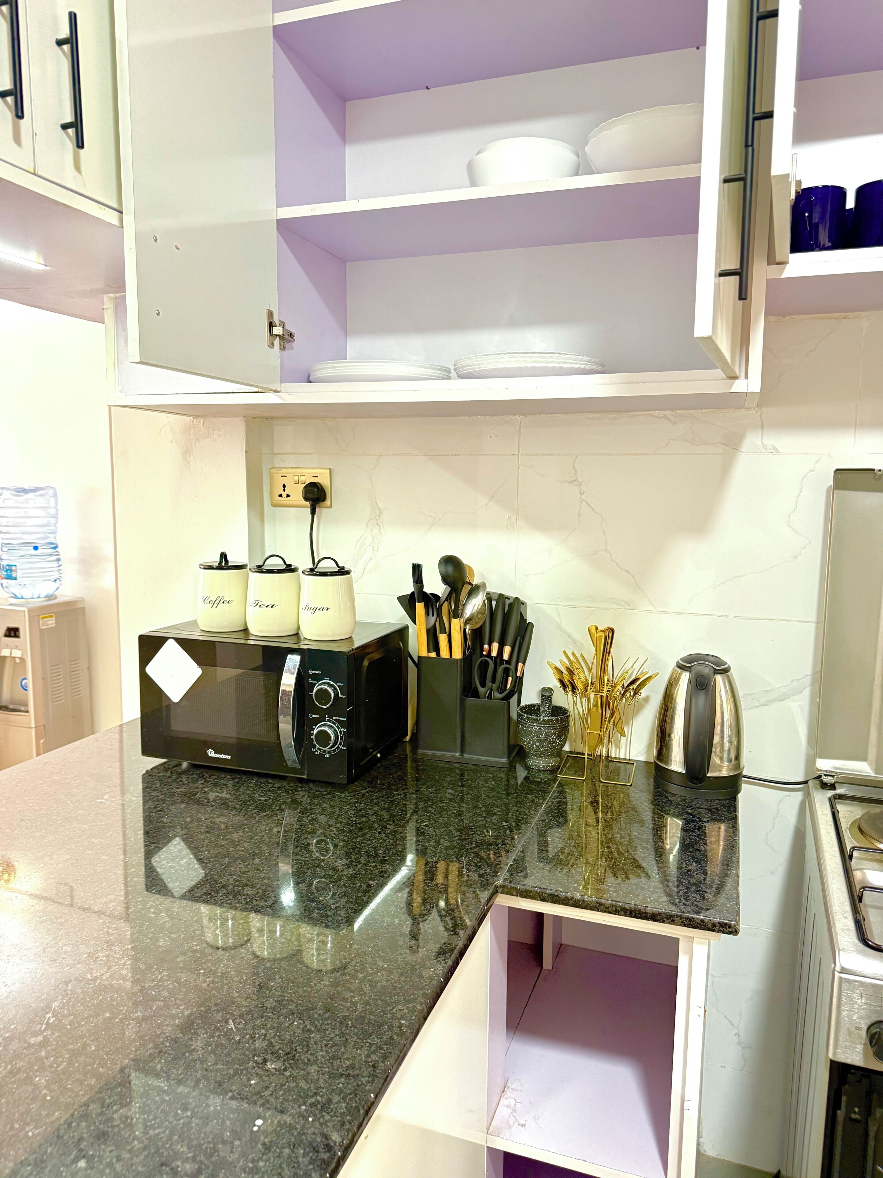 A kitchen with a microwave, toaster, and kettle on a granite countertop. There are utensils and a water dispenser in the background.