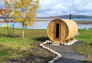 Una estructura de madera en forma de barril con una puerta, rodeada por un camino de grava y un pequeño borde de piedra, con un lago y árboles al fondo.