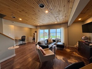 Ein großzügiger Wohnbereich mit Holzboden, einem großen Fenster mit Blick ins Freie, einem Ledersofa und einem an der Wand befestigten Fernseher.