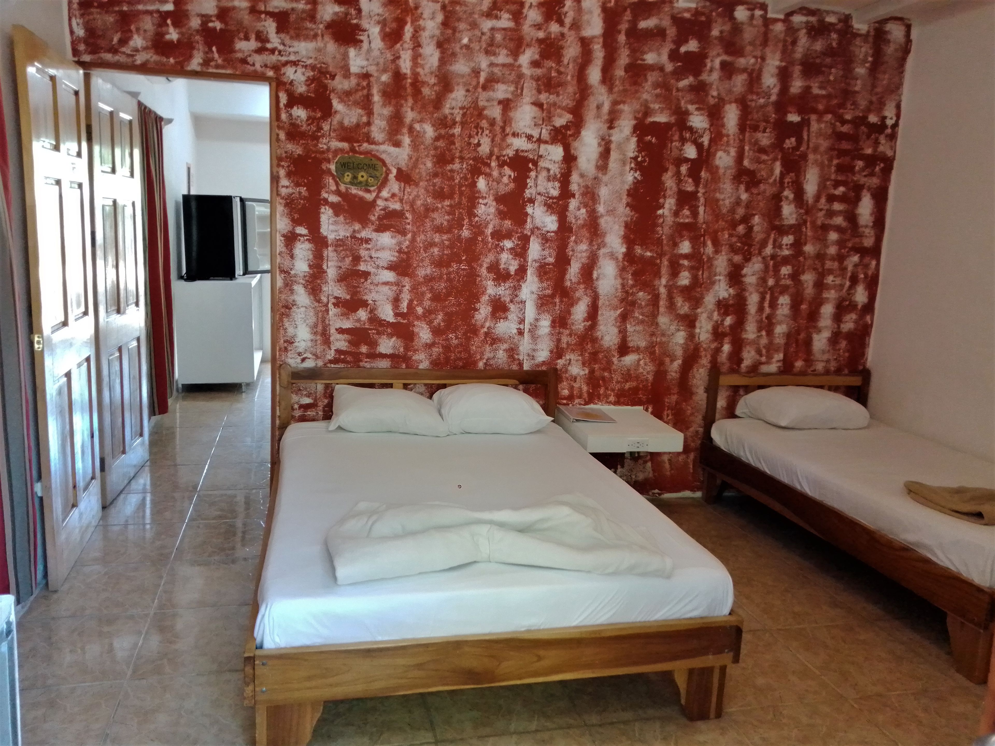 Una habitación con una cama grande, una cama más pequeña, una mesita de noche y un televisor montado en la pared.