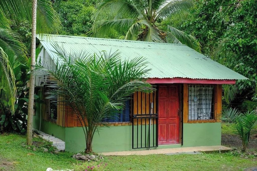 A small, single-story building with a green exterior and a red door, surrounded by lush greenery.