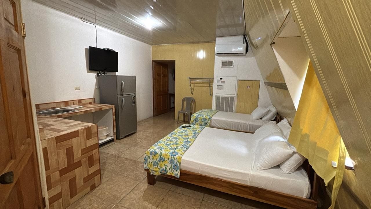 A compact room with a kitchenette, a bed, a television, and an air conditioning unit.