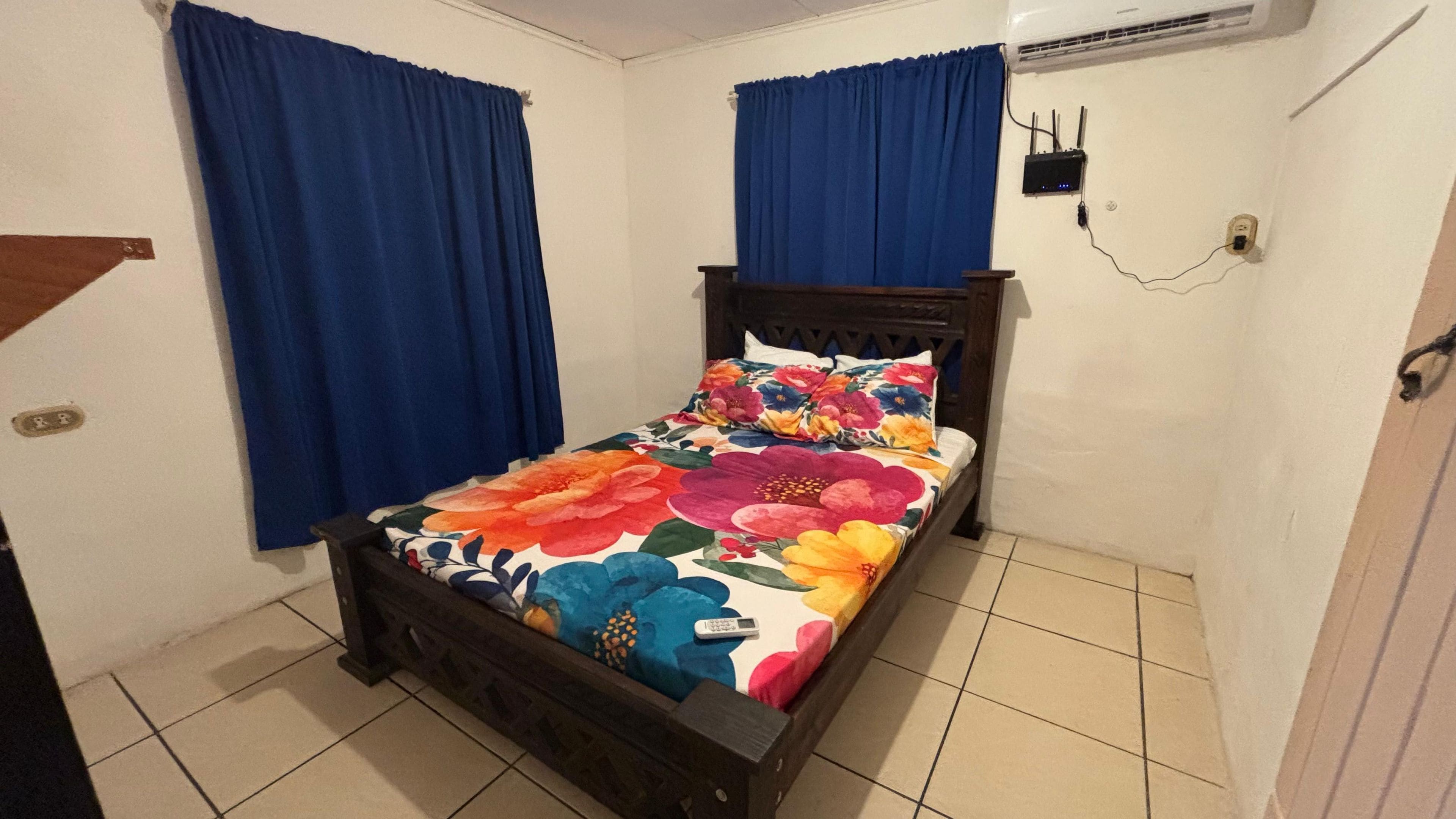 Une chambre à coucher avec un lit, des rideaux bleus, une tête de lit en bois, un mur blanc, un sol carrelé et un climatiseur fixé au mur.