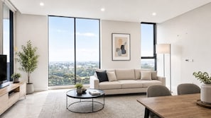 Una sala de estar moderna con un sofá, una mesa de centro y un televisor sobre un mueble de madera. Se ve una ventana grande con vistas a la ciudad.