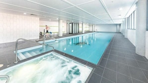 Ein großes Hallenbad mit separatem Whirlpoolbereich, umgeben von gefliestem Boden und Wänden sowie einer Glasdecke.