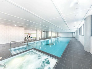 Una amplia piscina interior con un área de hidromasaje separada, rodeada de piso y paredes con azulejos, y un techo de vidrio.