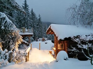 En snedækket træhytte med sne på taget og en træterrasse.