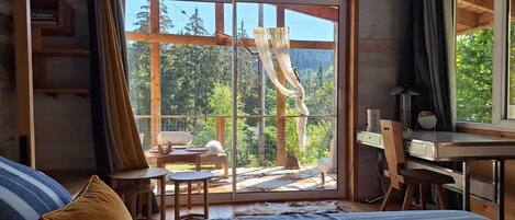 A bedroom with a large sliding glass door leading to a balcony, a bed with a striped cover, a desk with a chair, and a small table with chairs.