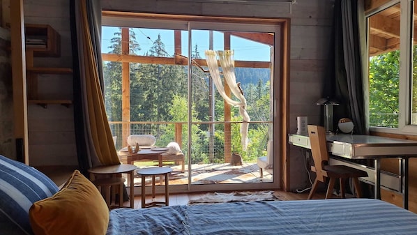 A bedroom with a large sliding glass door leading to a balcony, a bed with a striped cover, a desk with a chair, and a small table with chairs.