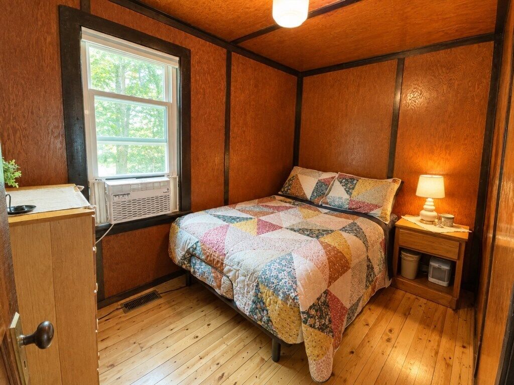 Un dormitorio con piso de madera, una cama con colcha colorida, una mesita de noche con lámpara y una ventana con persianas.