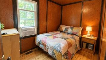 Un dormitorio con piso de madera, una cama con colcha colorida, una mesita de noche con lámpara y una ventana con persianas.