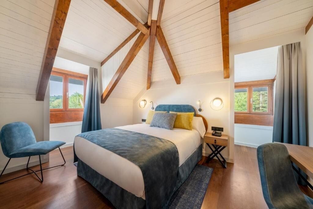 Un dormitorio con una cama grande, vigas de madera, dos mesitas de noche y vistas a la vegetación por las ventanas.