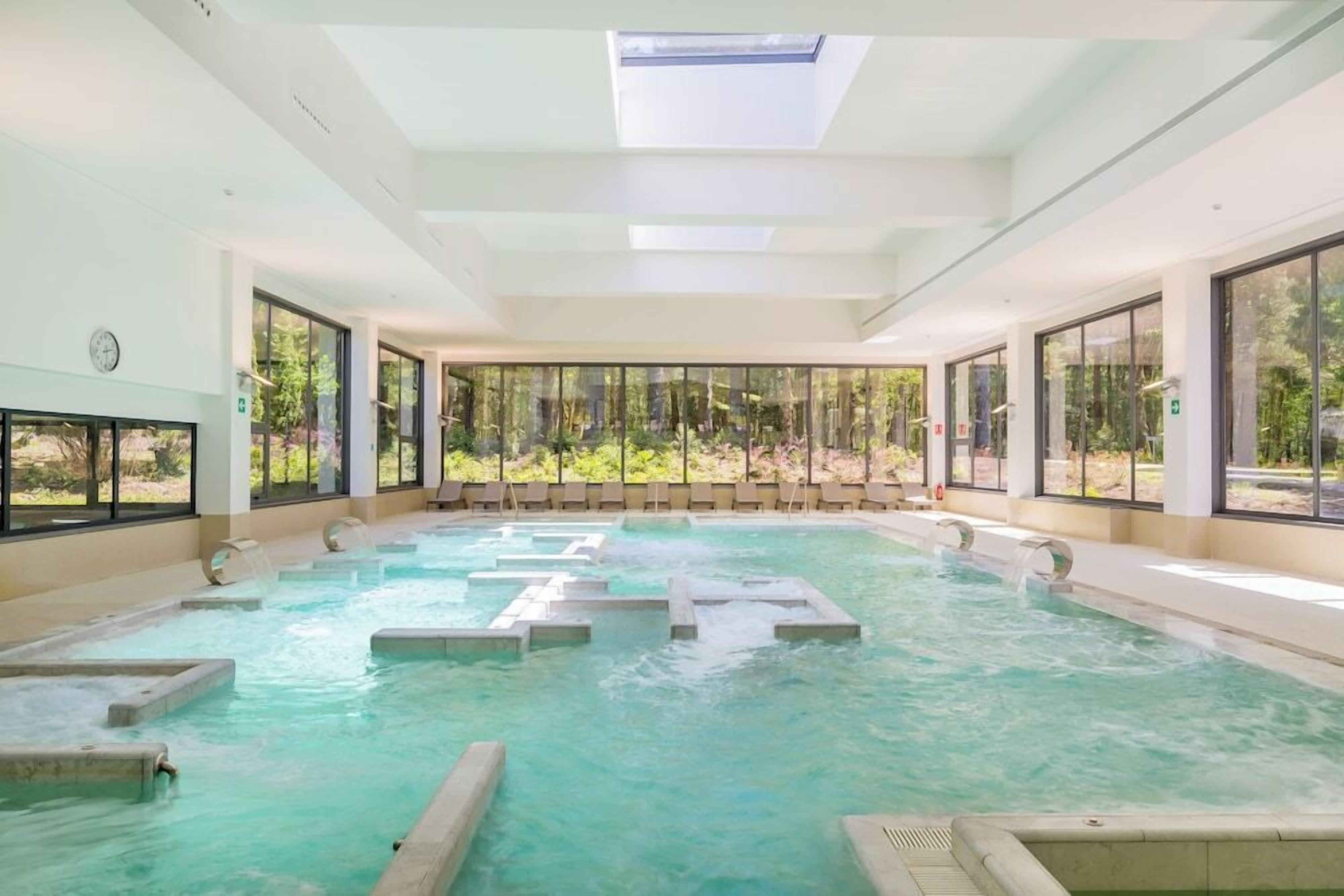 A large indoor swimming pool with clear water, surrounded by large windows offering views of greenery.