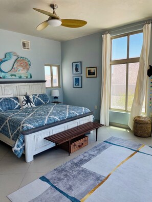 Un dormitorio con cama, un banco, ventilador de techo, ventana con cortinas y decoración en la pared.