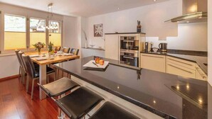Une cuisine moderne dotée d’un îlot central, d’un coin repas avec des chaises et offrant une vue sur l’extérieur grâce à de grandes fenêtres.