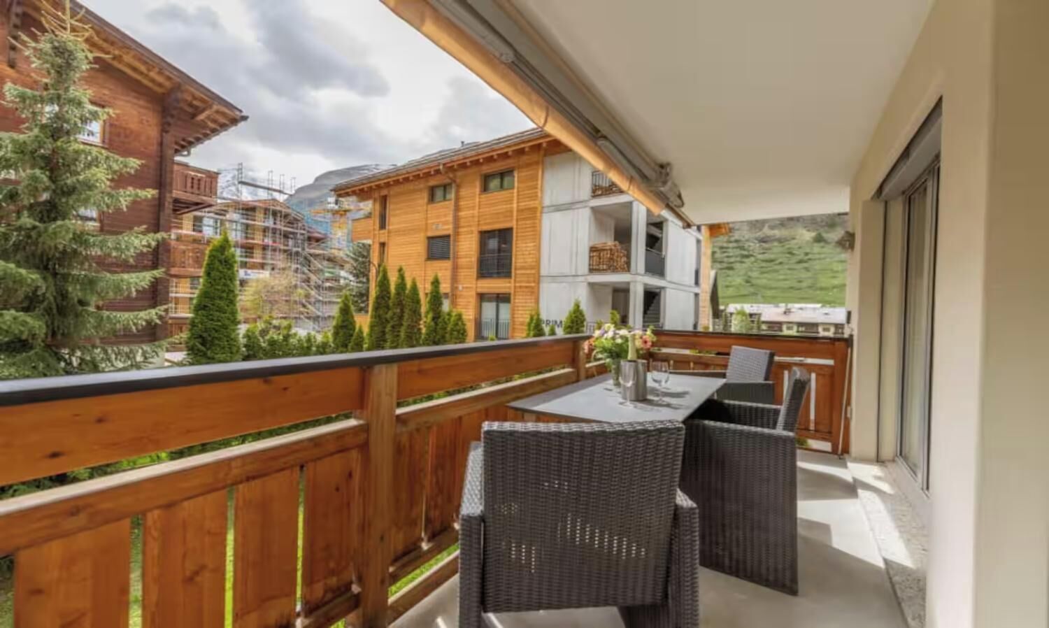 Un balcon agrémenté de meubles en osier, une table avec un vase, et une vue sur les bâtiments et les arbres.