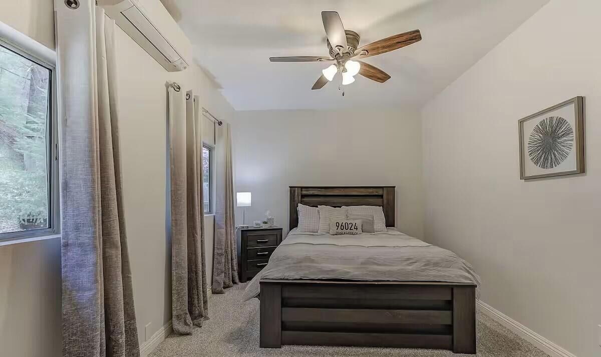 Un dormitorio con cama, mesita de noche, ventilador de techo y ventana con cortinas.