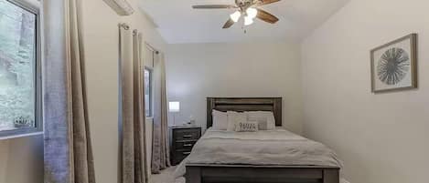 Un dormitorio con cama, mesita de noche, ventilador de techo y ventana con cortinas.