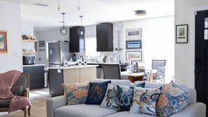 Uma sala de estar moderna com um sofá cinzento, almofadas estampadas, uma mesa de centro de vidro e uma cozinha ao fundo.