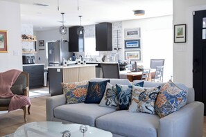 Ein modernes Wohnzimmer mit grauem Sofa, gemusterten Kissen, einem Glastisch und einer Küche im Hintergrund.