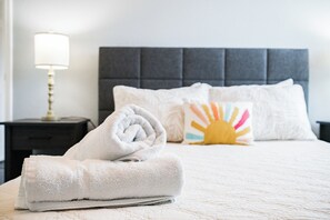 Un lit bien fait, avec une serviette blanche roulée, un oreiller orné d’un motif solaire et une lampe de chevet posée sur une table de nuit.