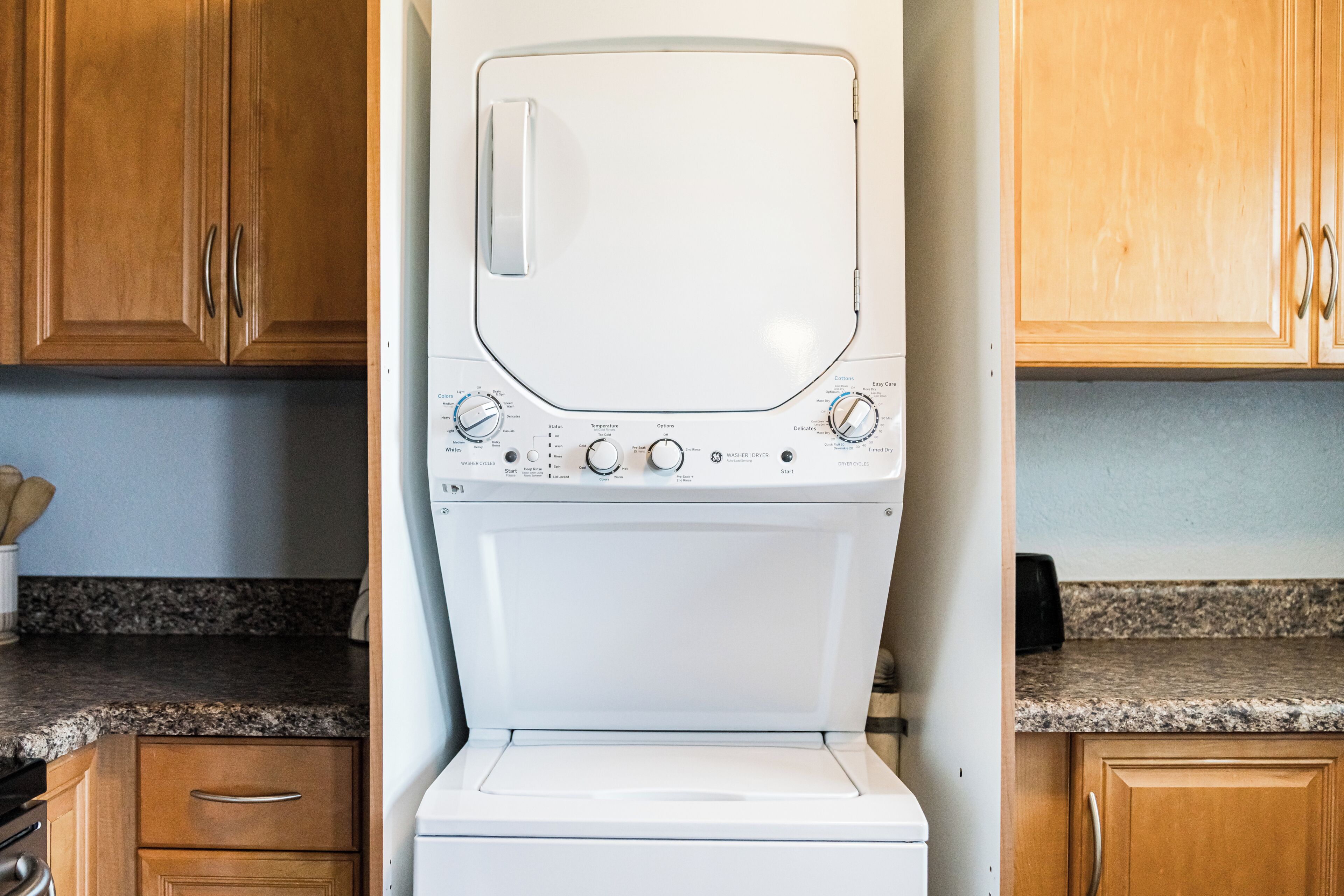 A front view of a white washing machine with control knobs and buttons, set in a kitchen with wooden cabinets and a granite countertop.