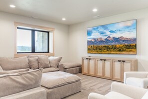 A modern living room with a large window, a sofa set, a flat-screen TV mounted on a wooden cabinet, and a mountain landscape painting on the wall. - The Lodge at Beach Meadow | Hot Tub + Pickleball (Island Park)
