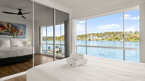 A hotel room with a bed, a painting, a ceiling fan, and a large window with a view of a lake and houses.