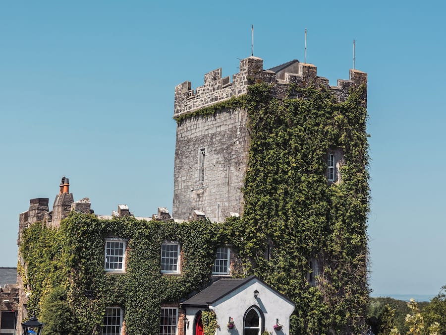 Um edifício imponente com estilo de castelo, apresentando uma torre notável, paredes cobertas de hera e uma pequena construção adjacente com uma porta vermelha.