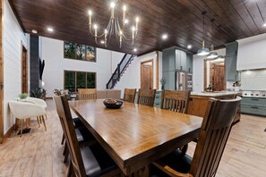 A dining area with a wooden table and chairs, a chandelier, and a kitchen in the background. - 5BR Hochatown Cabin: Hot Tub - Game Room - Fits 13 - Min to Lake & Casino (Broken Bow)