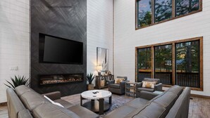 A modern living room with a fireplace, comfortable seating, and large windows. - 5BR Hochatown Cabin: Hot Tub - Game Room - Fits 13 - Min to Lake & Casino (Broken Bow)
