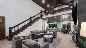 A spacious living area with a staircase, a sofa set, a coffee table, and a kitchen in the background. - 5BR Hochatown Cabin: Hot Tub - Game Room - Fits 13 - Min to Lake & Casino (Broken Bow)