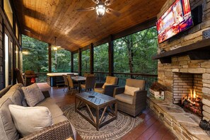 A covered outdoor seating area with a fireplace, wooden furniture, and a television screen. - Hochatown Luxury Cabin on 1+ Acre: Main Lodge + Detached Private Suite & Stream (Broken Bow)