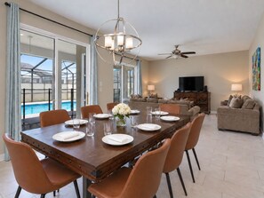 A dining area with a large table, chairs, and a chandelier. There is a pool visible through the glass doors. - Walk to Lazy River - Themed 6BR Home - Private Pool, Spa and Game Room, Store... (Kissimmee)