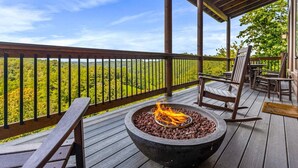 Een houten terras met vuurkorf, buitenzitplaatsen en uitzicht op een bebost landschap.