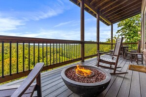 Eine Holzterrasse mit Feuerstelle, Sitzgelegenheiten im Freien und Blick auf eine bewaldete Landschaft.