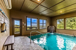 Ein Hallenbad mit Steinboden, einer Metallleiter und Blick auf eine natürliche Landschaft durch große Fenster.