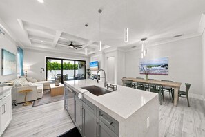 A modern kitchen with a central island, a dining area with a long table, and a living space with a sofa and coffee table. - New! Coral Tides + Brand 5BR New Luxury Build + Heated Pool + 1 Mile to Vanderbilt Beach! (Naples)
