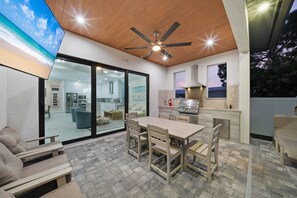 An outdoor kitchen area with a dining table and chairs, a ceiling fan, and a view of the interior through a glass door. - New! Coral Tides + Brand 5BR New Luxury Build + Heated Pool + 1 Mile to Vanderbilt Beach! (Naples)
