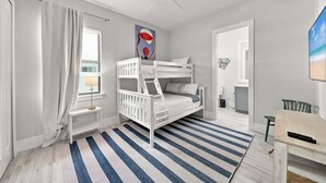 Een kinderkamer met stapelbedden, een plafondventilator, een raam met gordijnen, een schilderij en een strandafbeelding op de muur.