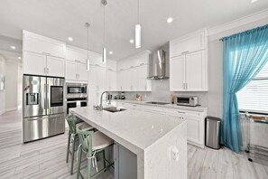 A modern kitchen with white cabinetry, stainless steel appliances, a marble countertop, and a central island with bar stools. - New! Coral Tides + Brand 5BR New Luxury Build + Heated Pool + 1 Mile to Vanderbilt Beach! (Naples)