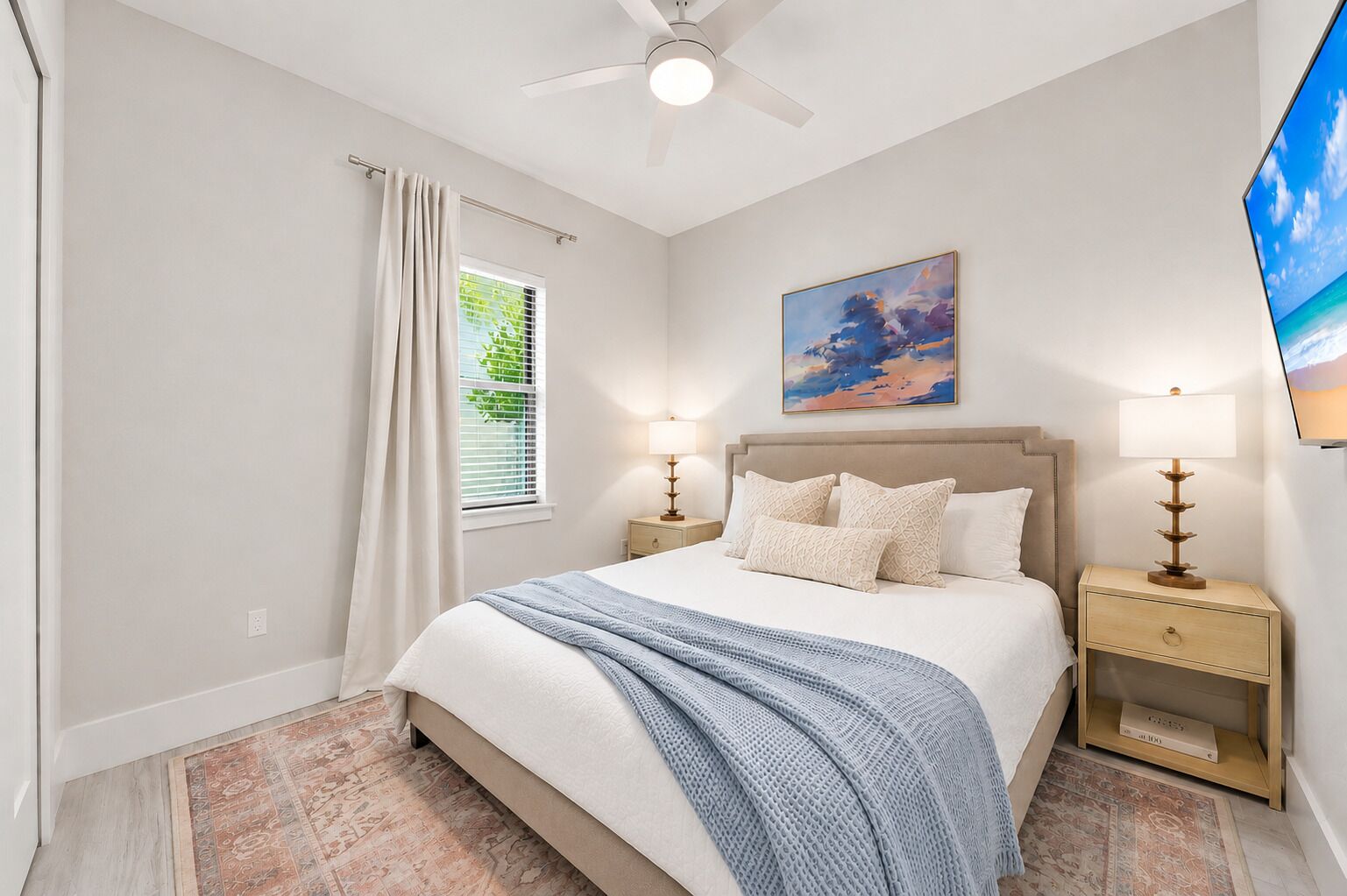 A bedroom with a bed, bedside tables, a ceiling fan, a window with curtains, a painting, and a television.