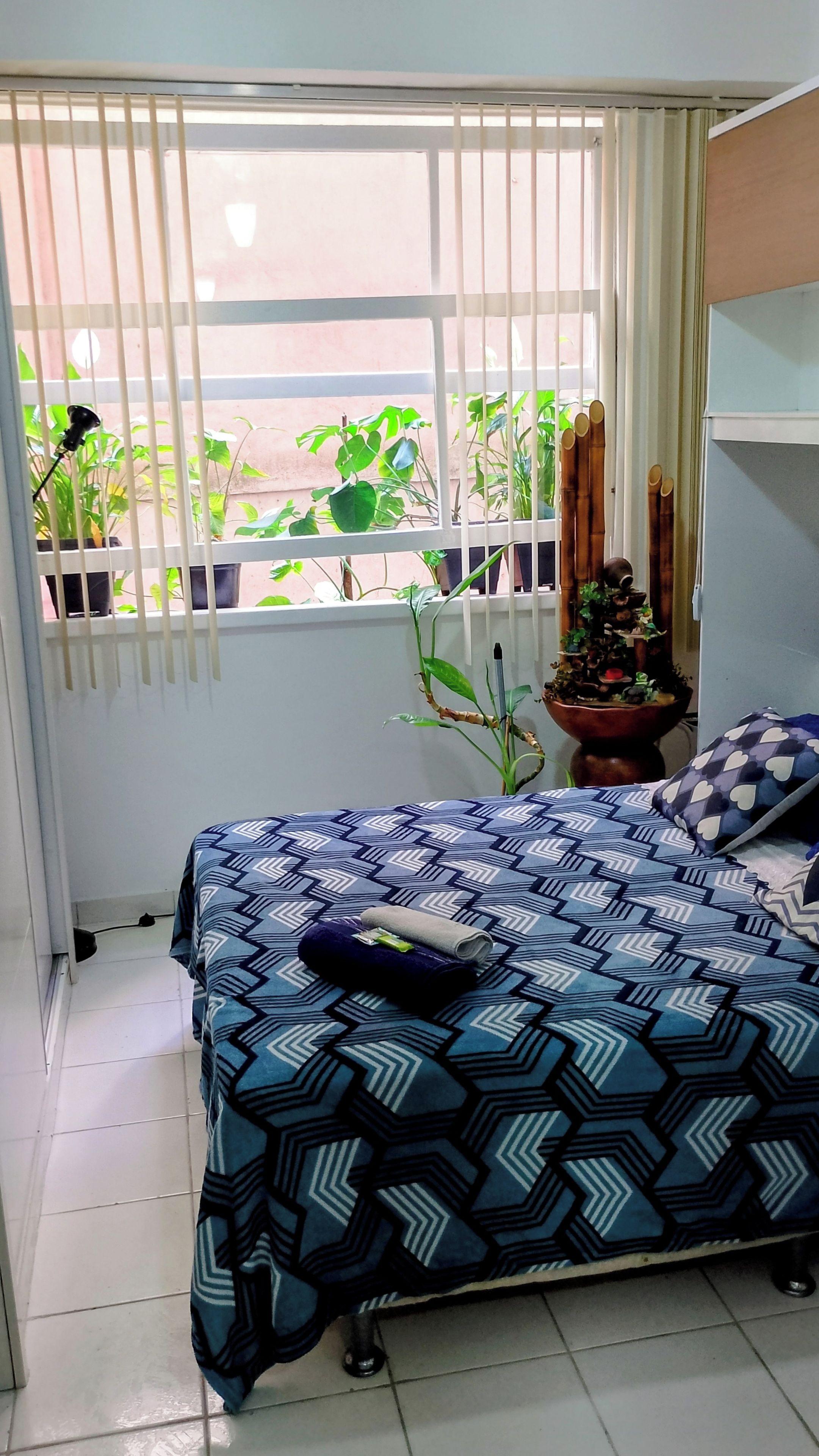 A bedroom with a bed, a window with blinds, a potted plant, and a bedside table.