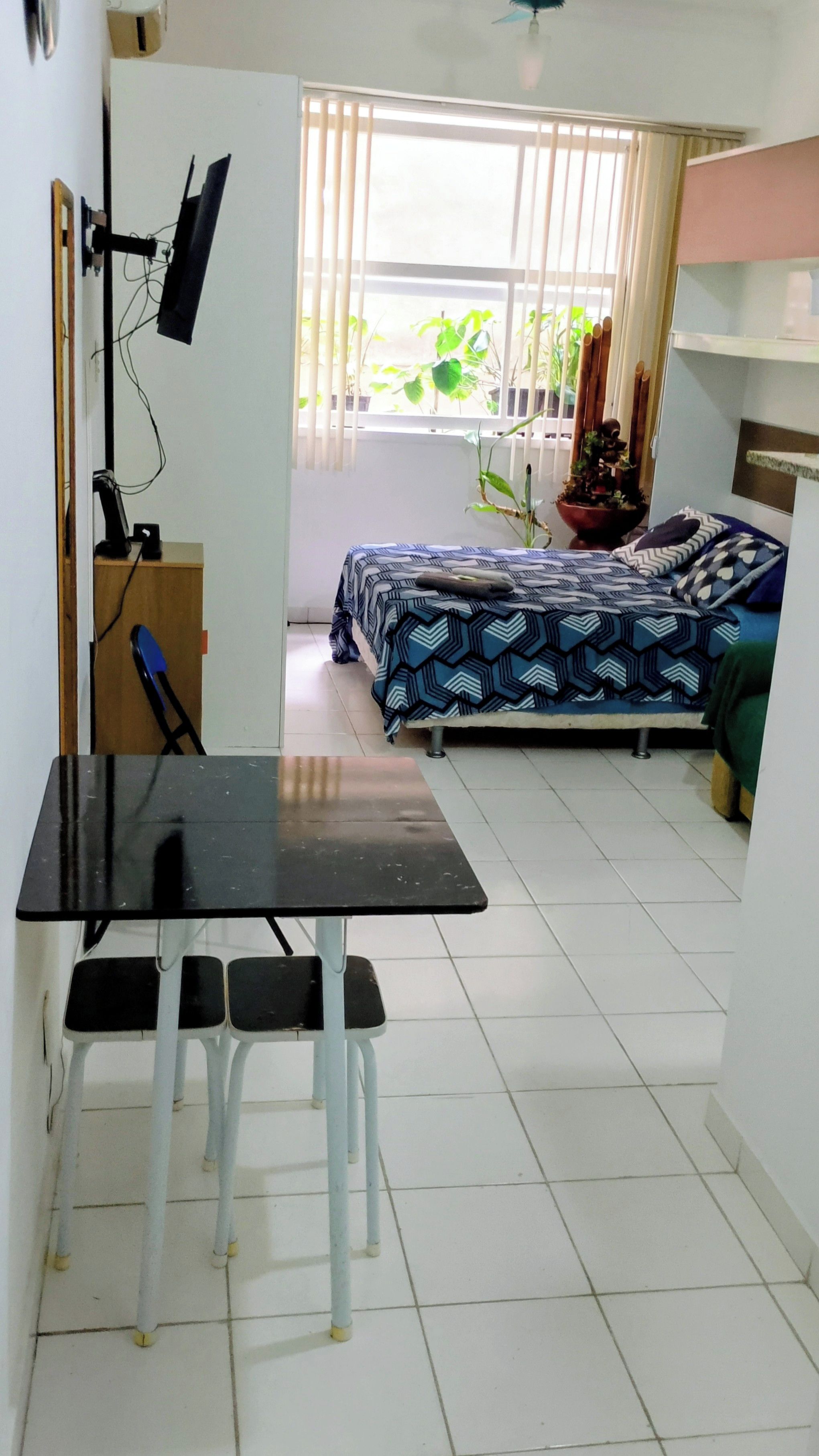 Ein Zimmer mit einem Bett, einem Schreibtisch mit Stuhl, einem Fernseher und einem Fenster mit Jalousien.