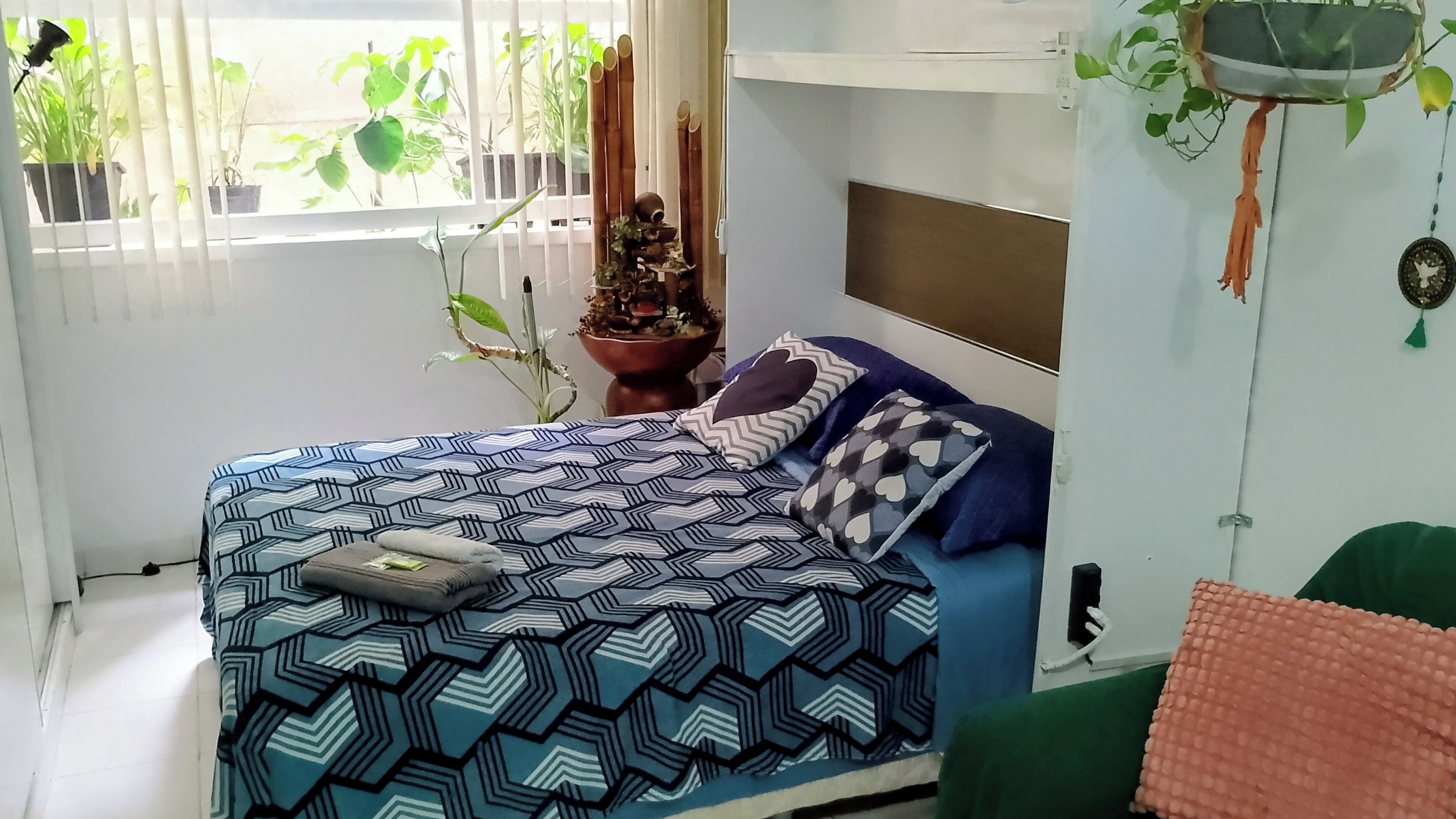 A bed with a patterned bedspread, a nightstand with a decorative item, and a window with plants.