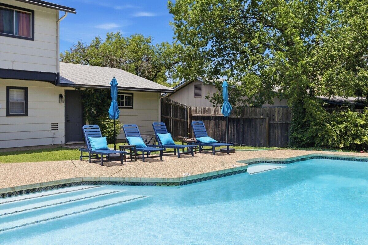 Una piscina con sillones de descanso y sombrillas, rodeada por una valla de madera y árboles.