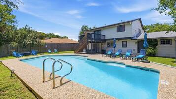 A rectangular swimming pool with a pebble-lined edge, surrounded by a wooden deck. There are lounge chairs and a covered patio area with umbrellas.
