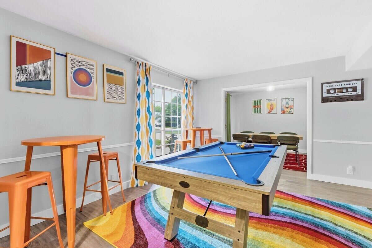 A pool table in a room with a colorful striped carpet, orange stools, and framed artwork on the walls.